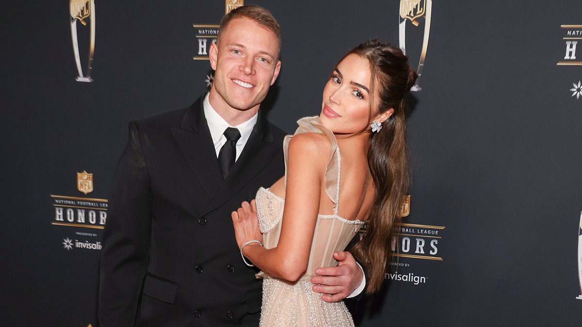 Christian McCaffrey smiles in a black suit as Olivia Culpo looks back at the cmaera and puts her hand on his chest