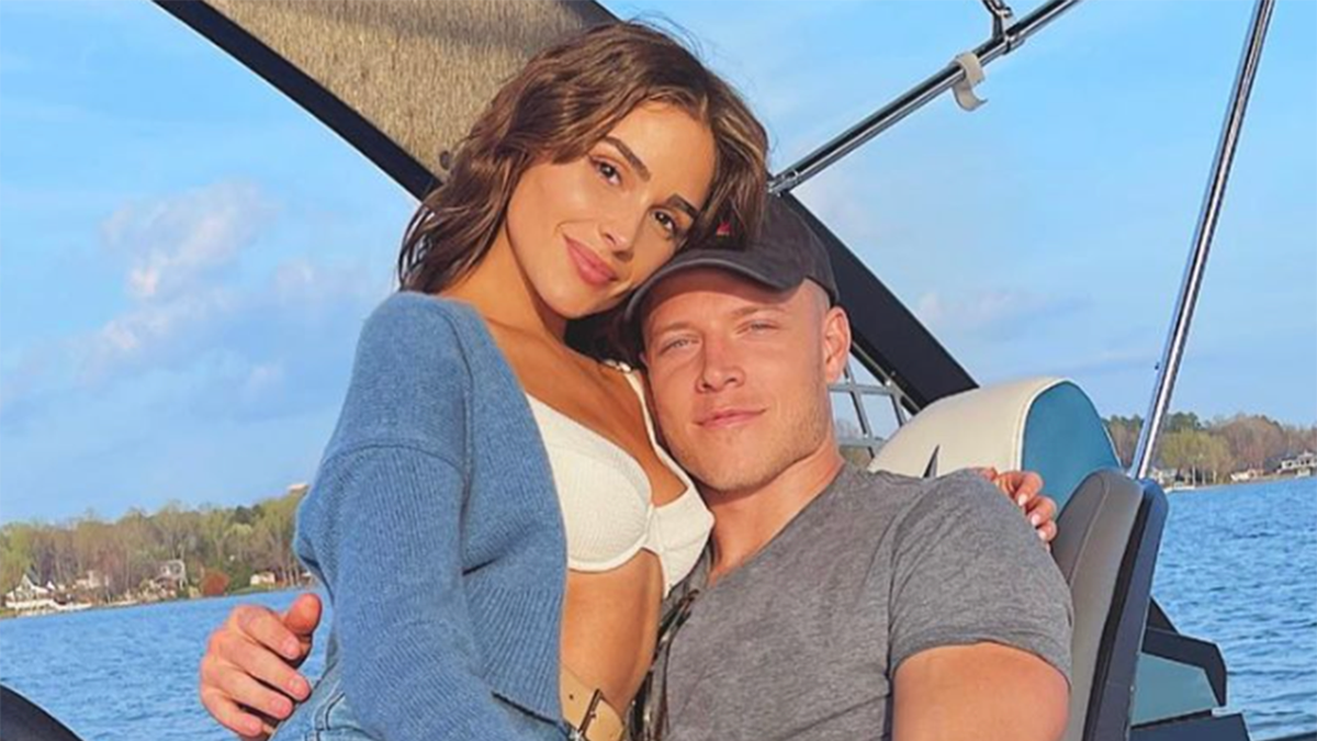 Olivia Culpo in a white bikini and blue sweater holds onto Christian McCaffrey in a grey shirt and black hat