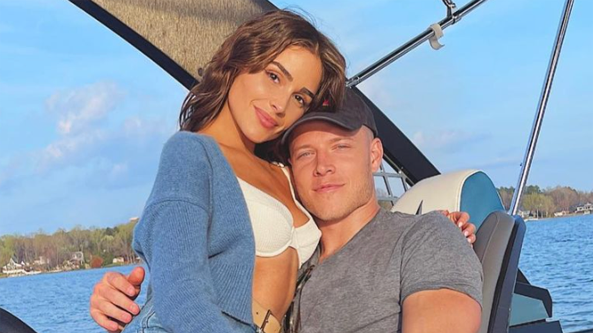 Olivia Culpo in bikini bianco e maglione blu si aggrappa a Christian McCaffrey con una camicia grigia e un cappello nero