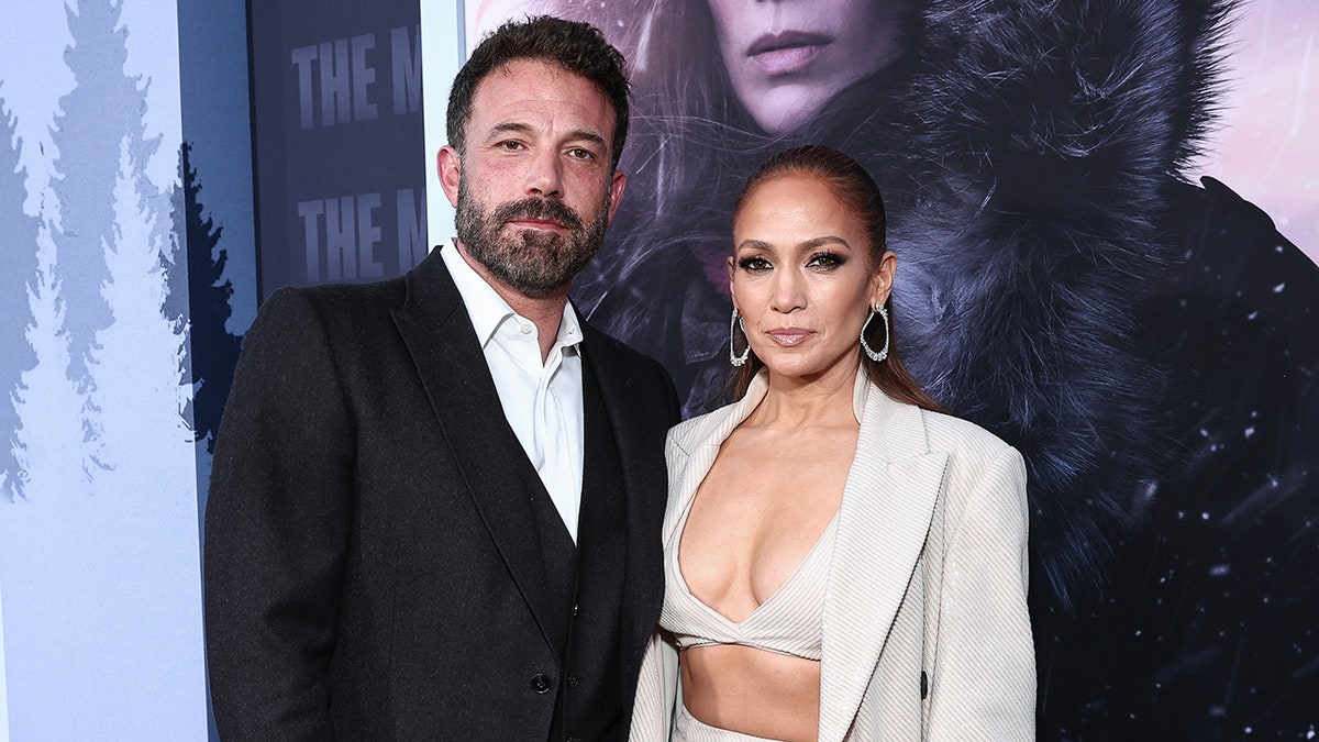 Ben Affleck in a black suit and white shirt poses on the carpet with wife Jennifer Lopez in a bra top and open blazer