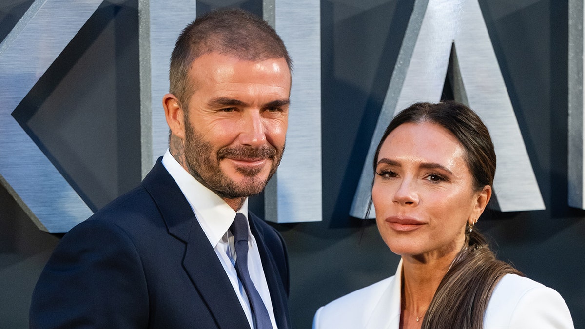 David Beckham in a blue suit and tie stands next to wife Victoria in a white suit on the "BECKHAM" carpet