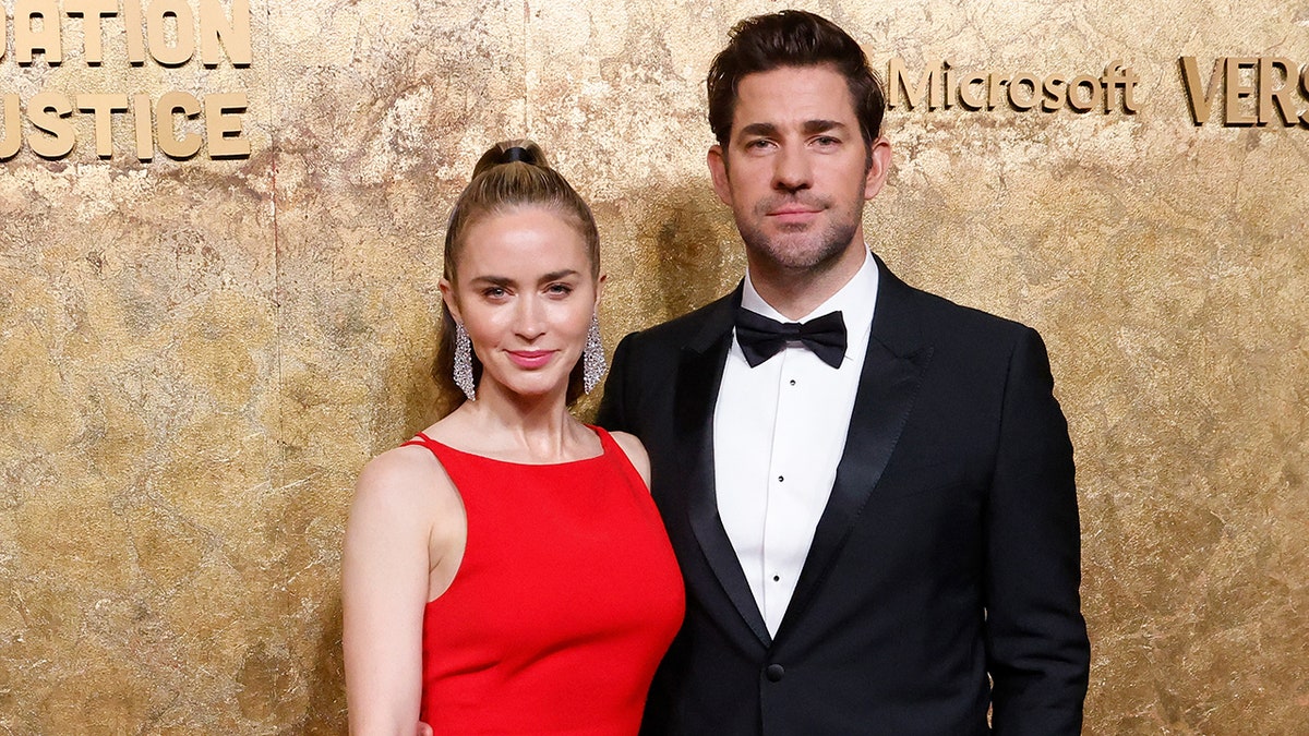 Emily Blunt in a red gown poses next to husband John Krasinski in a classic tuxedo on the carpet in New York City
