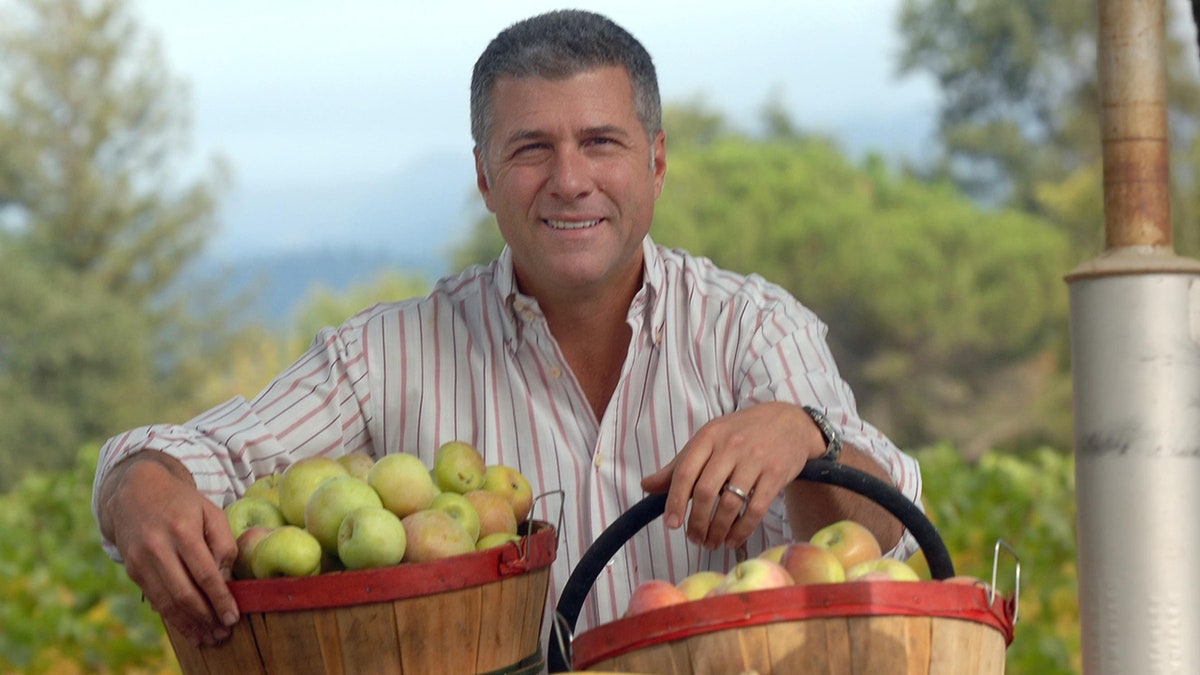 Michele Chiarello in camicia bianca a righe tiene cesti pieni di mele