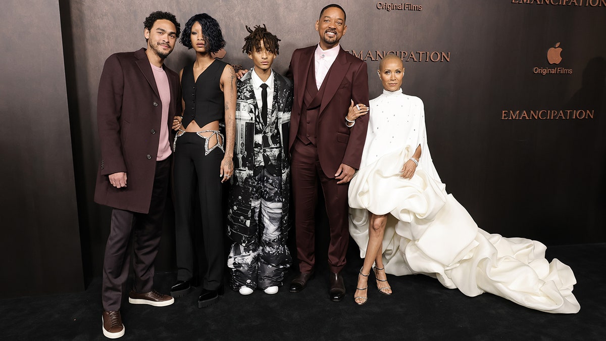 Will Smitt in a maroon suit poses with his three children and wife Jada Pinkett Smith on the carpet