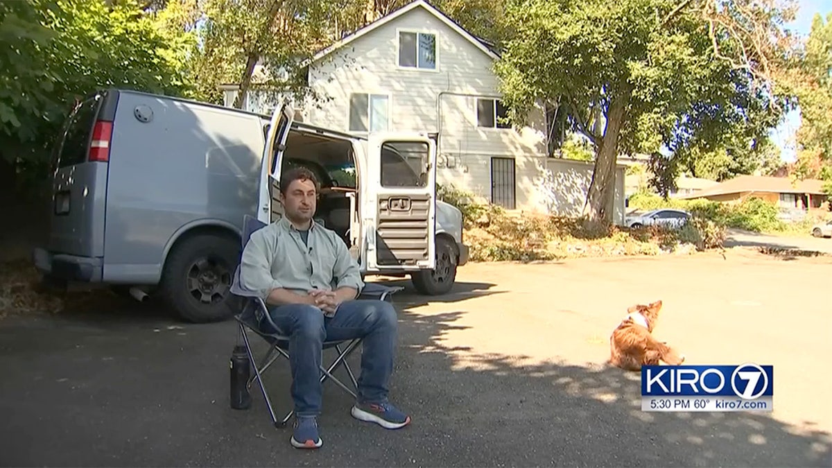 Man sits in lawn chair in front of white van with his house in background