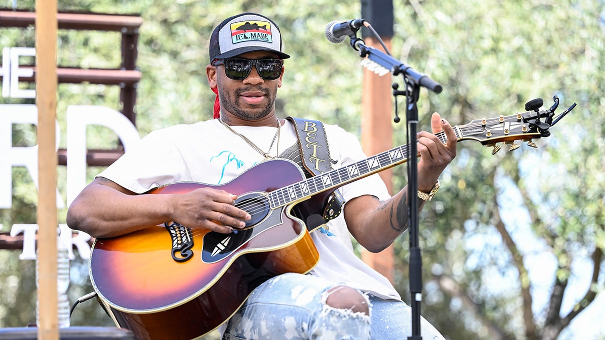 Jimmie Allen sits back in a white t-shirt and hat and strums the guitar on stage
