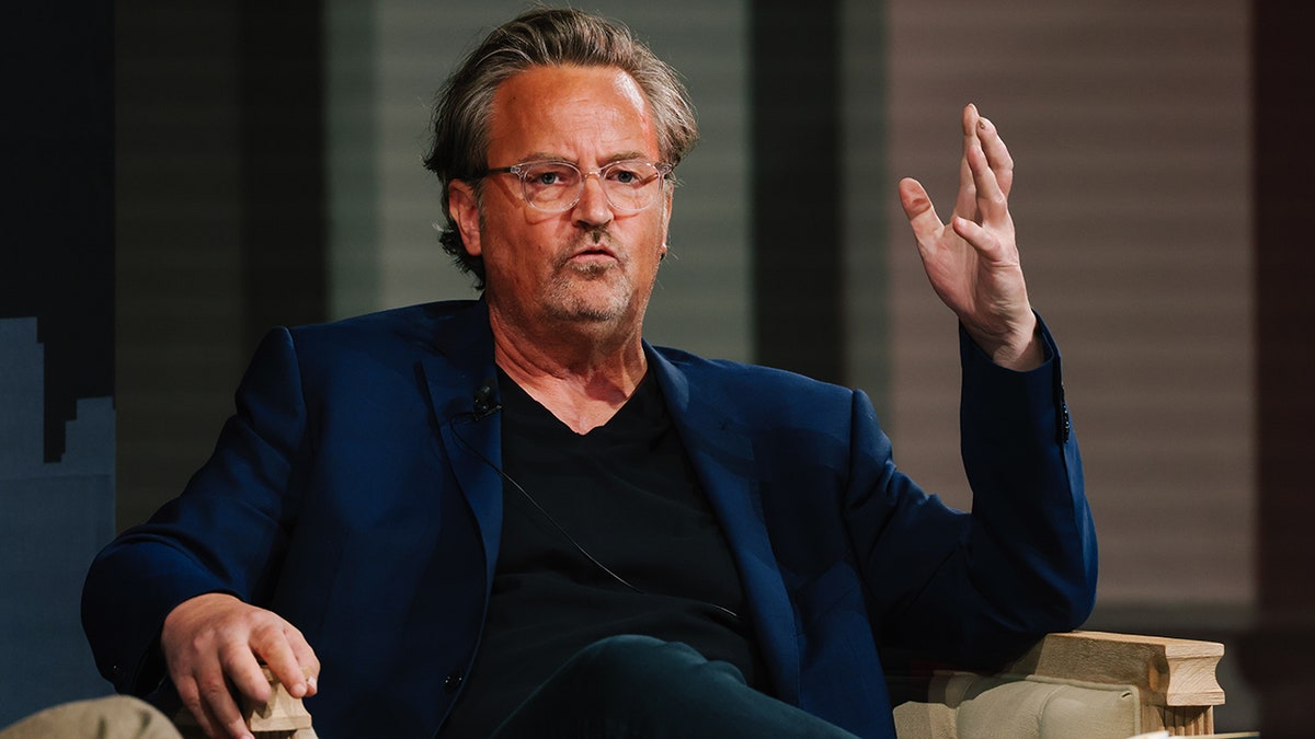 Matthew Perry sits down on stage in a blue suit and black shirt with his arm raised