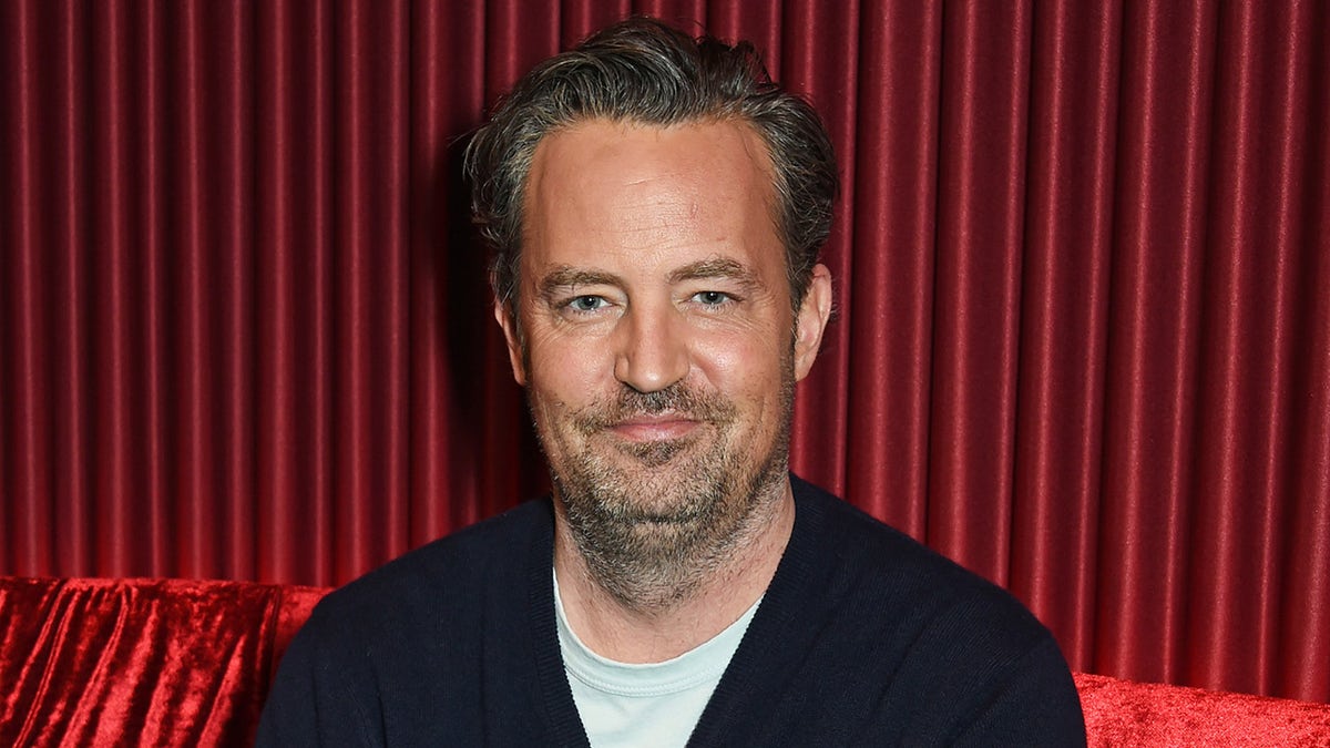 Matthew Perry w czarnym swetrze uśmiecha się delikatnie przed czerwoną zasłoną