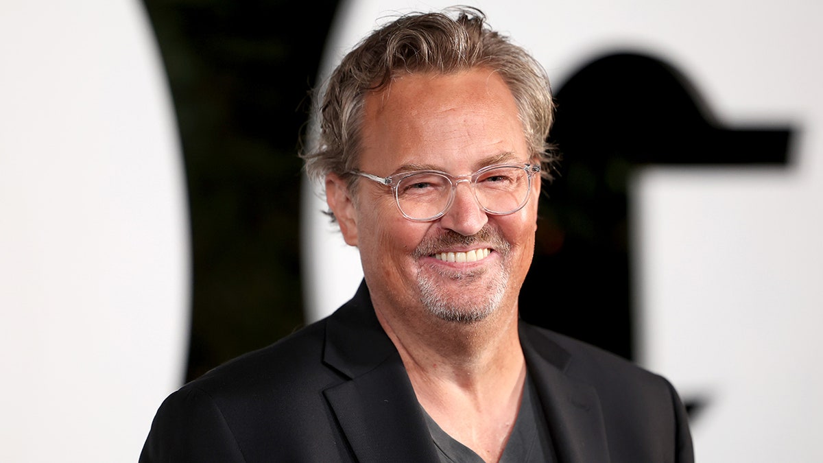 Matthew Perry smiles in a black jacket and shirt with clear glasses on the carpet