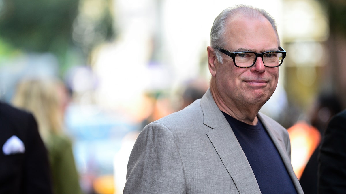 Glenn Gordon Caron looks at the camera with a grimace while walking outside in New York City, wearing a grey blazer and navy shirt and black square-framed glasses