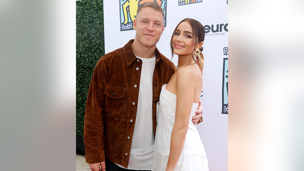 Christian McCaffrey in a brown jacket and white t-shirt smiles next to Olivia Culpo in a white dress