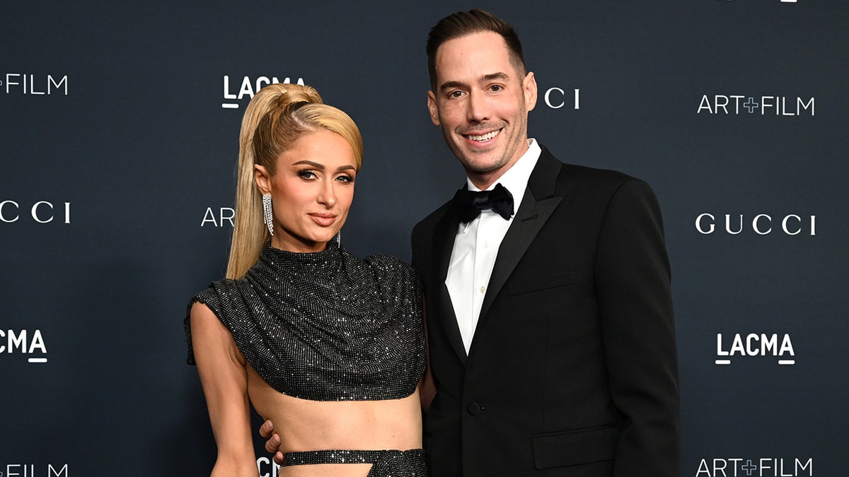 Paris Hilton in a sparkly cutout dress with pulled back hair poses with husband Carter Reum in a classic tuxedo on the carpet