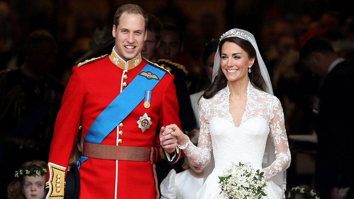 Prince William in a red uniform and blue sash holds Kate Middleton's hand in her wedding dress