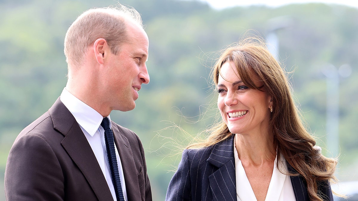 Prince William in a dark brown suit smiles at Kate Middleton in a navy suit in Wales