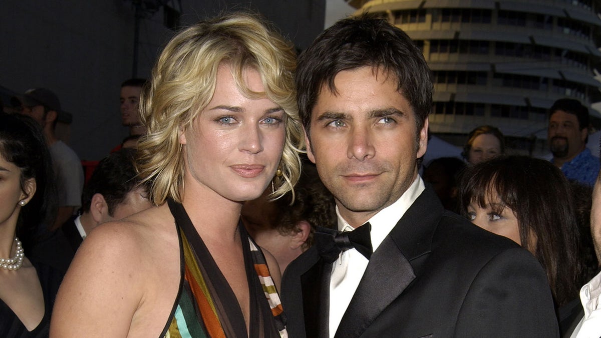 Rebecca Romijn in a striped halter top smiles next to John Stamos in a classic tuxedo