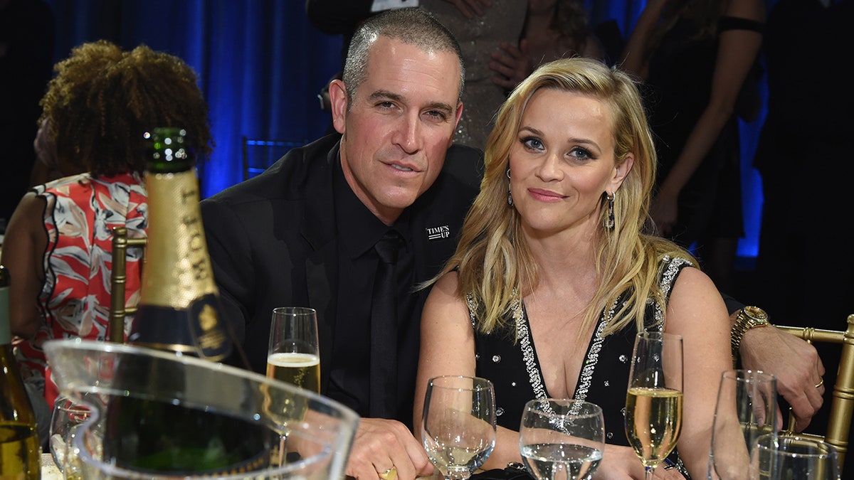 Jim Toth de terno preto e gravata posa em uma mesa com Reese Witherspoon em um vestido preto no Critics' Choice Awards