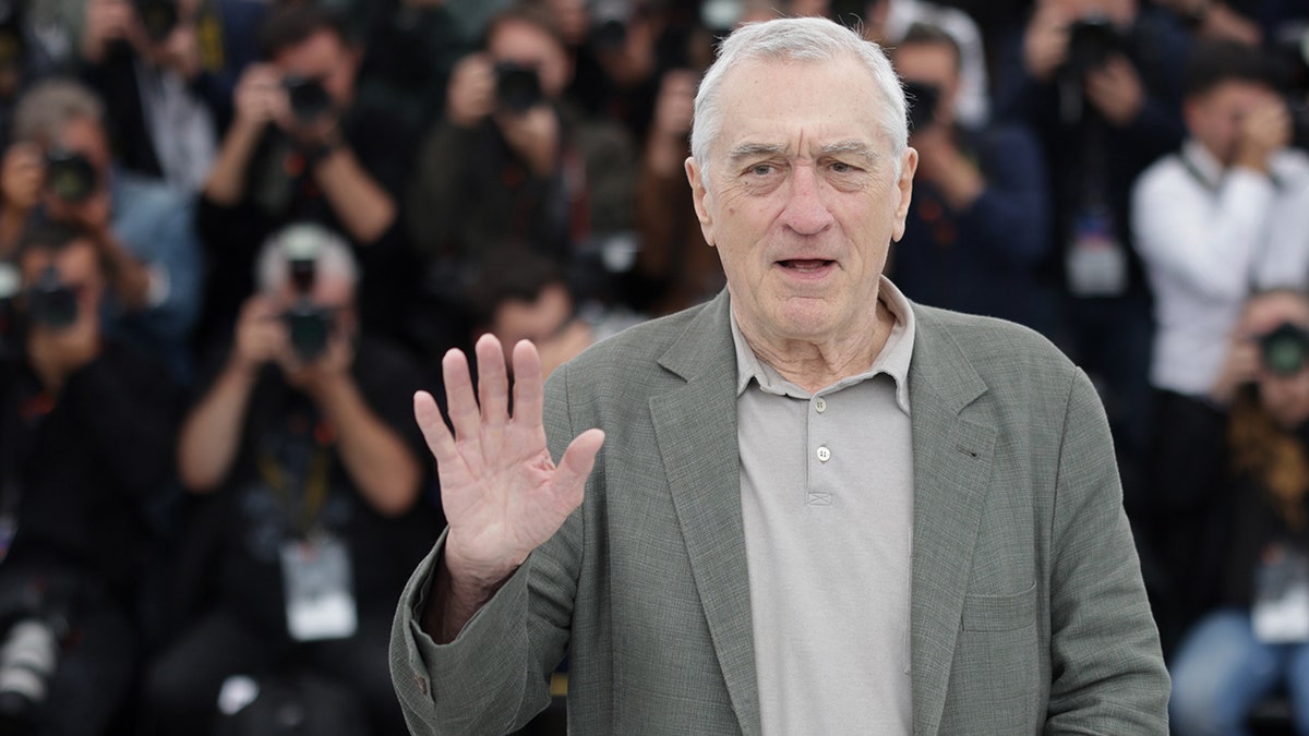 Robert De Niro in a grey suit puts up his right hand as he stands before the Cannes crowd