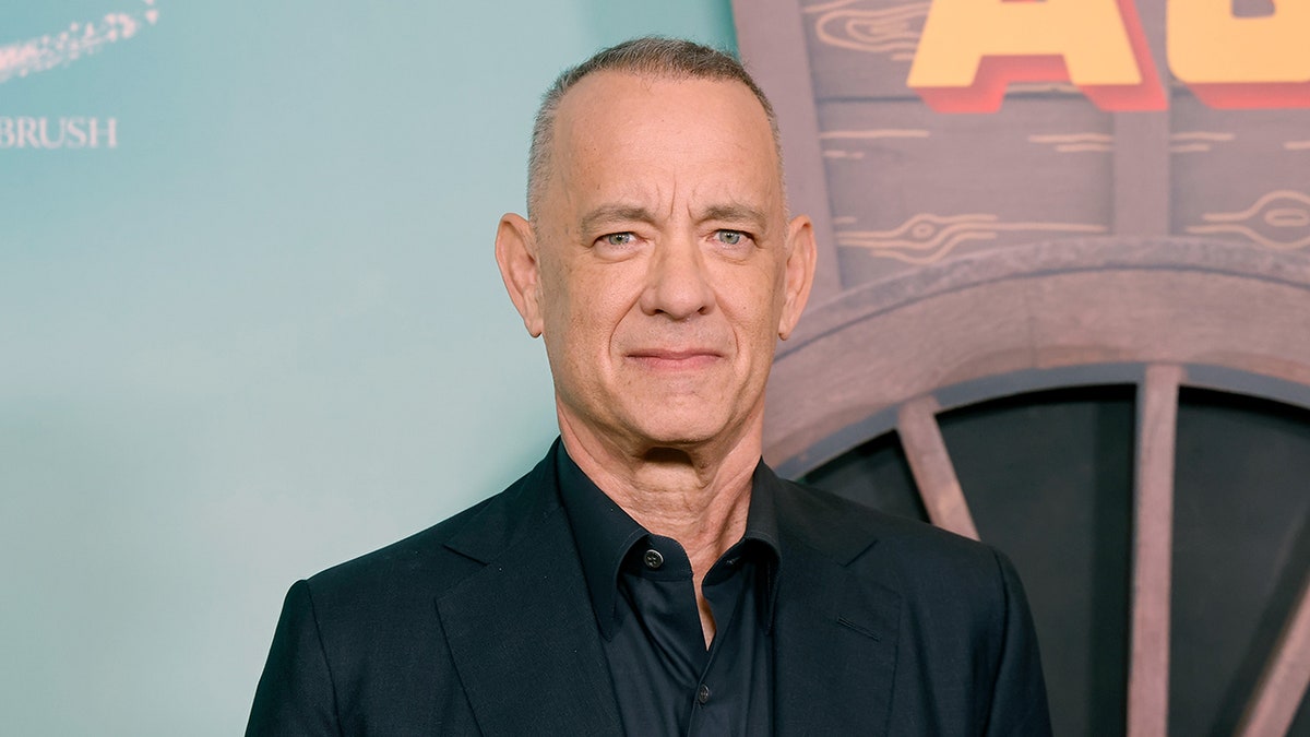 Tom Hanks appears stoic on the carpet in a dark suit and shirt.