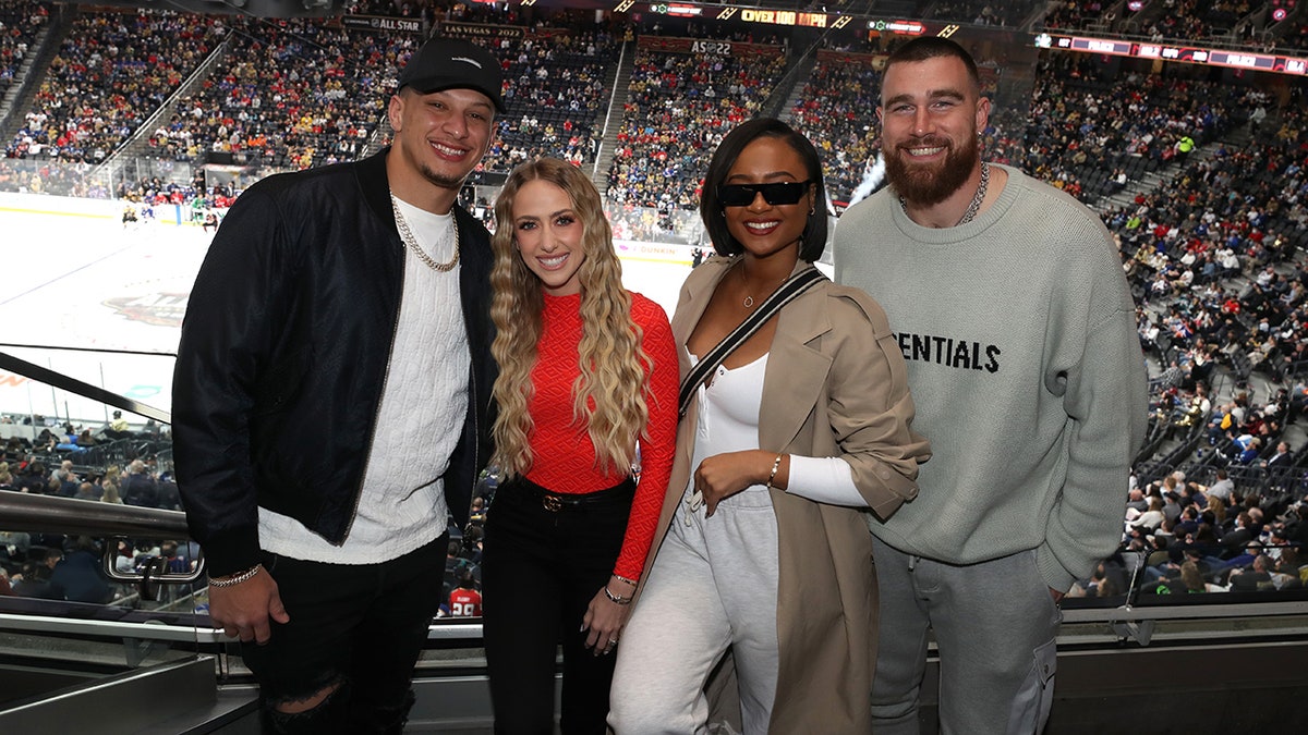 Patrick Mahomes in a white shirt and black jacket poses next to Brittany Mahomes in black pants and red turtleneck next to Kayla Nicole in all-white and tan trench coat next to Travis Kelce in a sweatshirt