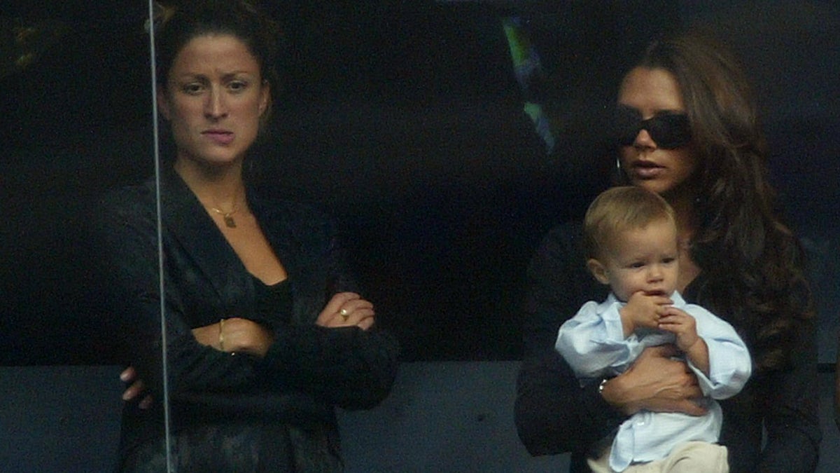 Rebecca Loos has her arms crossed in black standing in a suite next to Victoria Beckham in sunglasses and holding baby Romeo