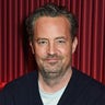 Matthew Perry sits in front of red curtains wearing blue sweater
