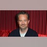 Matthew Perry in a black sweater soft smiles in front of red curtain