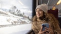 Portrait of a happy woman riding the train in the winter while using her phone and looking through the window