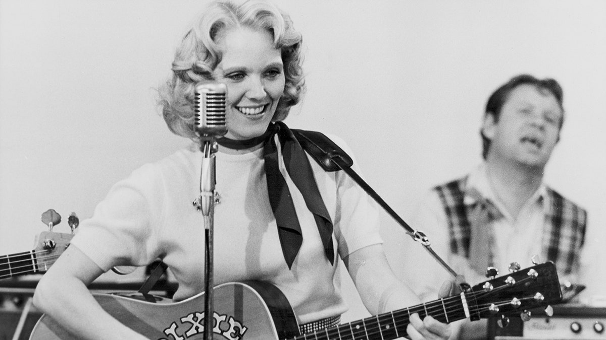 Conny Van Dyke holding guitar and smiling near a microphone