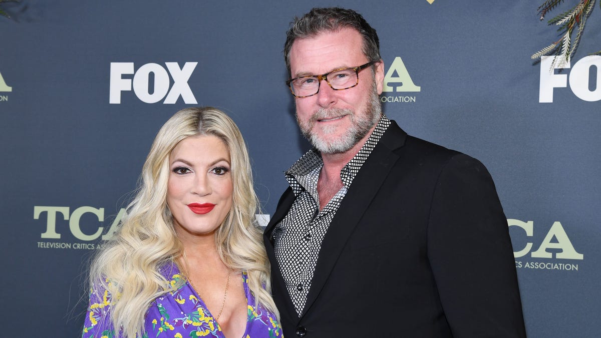 Tori Spelling in a purple blouse smiles with her husband Dean McDermott in a black jacket on the carpet