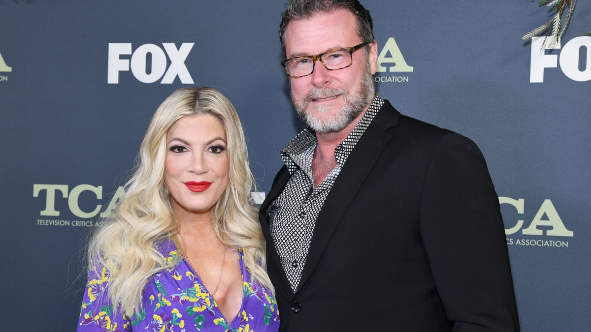 Tori Spelling in a purple blouse smiles with husband Dean McDermott in a black jacket on the carpet