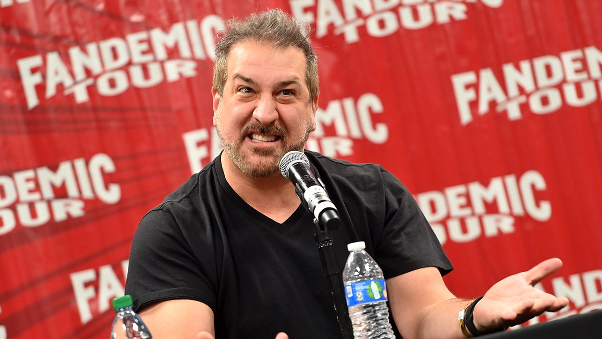 Joey Fatone in a black shirt sits behind a podium 