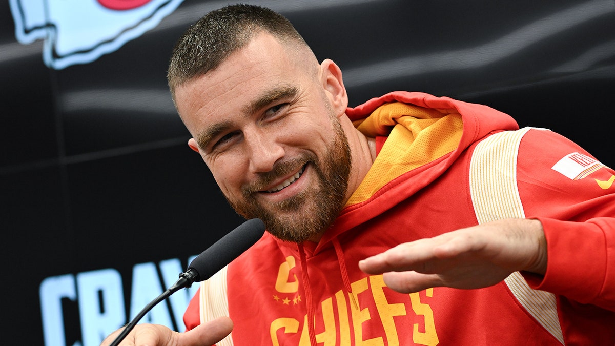 Travis Kelce in a red and yellow Chief's sweatshirt puts his hands in the air during a press conference