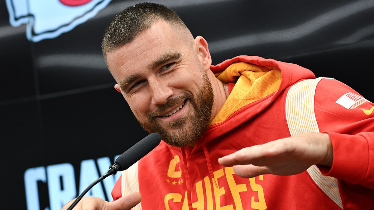 Travis Kelce con la felpa rossa e gialla del capo alza le mani in aria durante una conferenza stampa