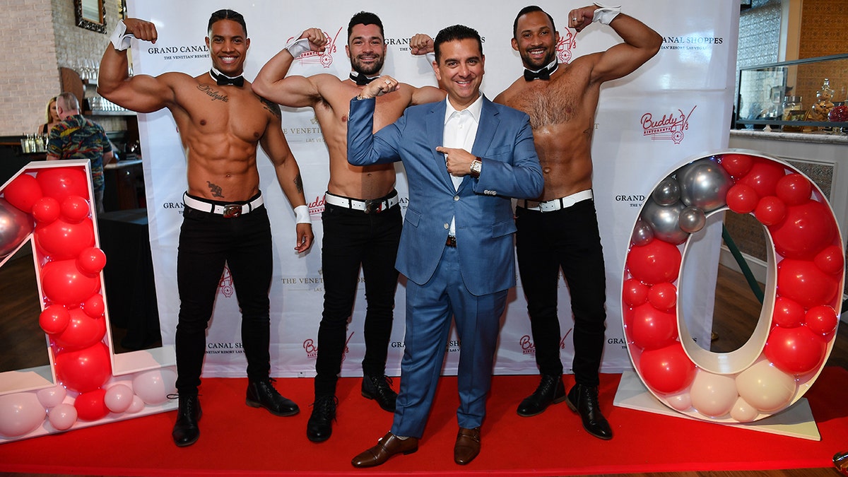 Buddy Valastro wearing a blue suit and a white shirt surrounded by shirtless male models