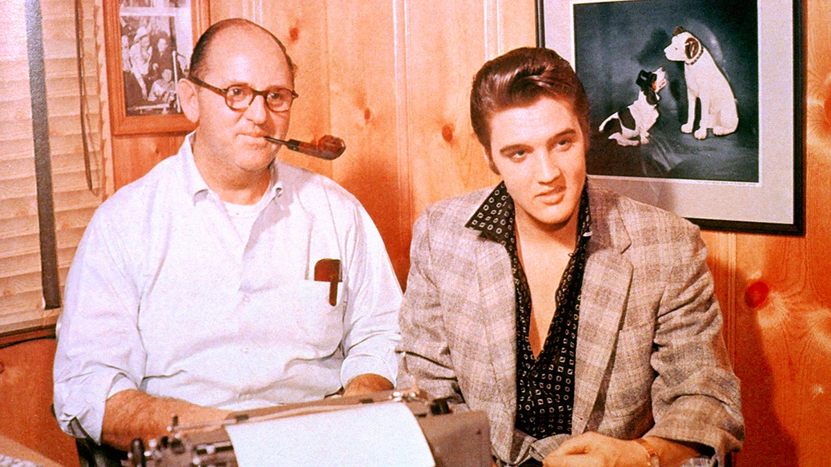 Elvis Presley em um blazer bege e uma camisa preta aberta ao lado de Tom Parker fumando um charuto