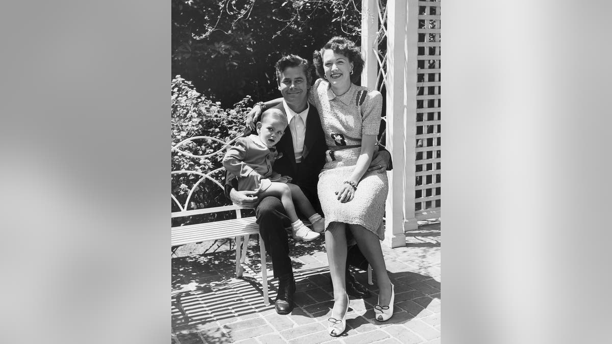 Eleanor Powell sentada no colo do marido enquanto ele segura o filho