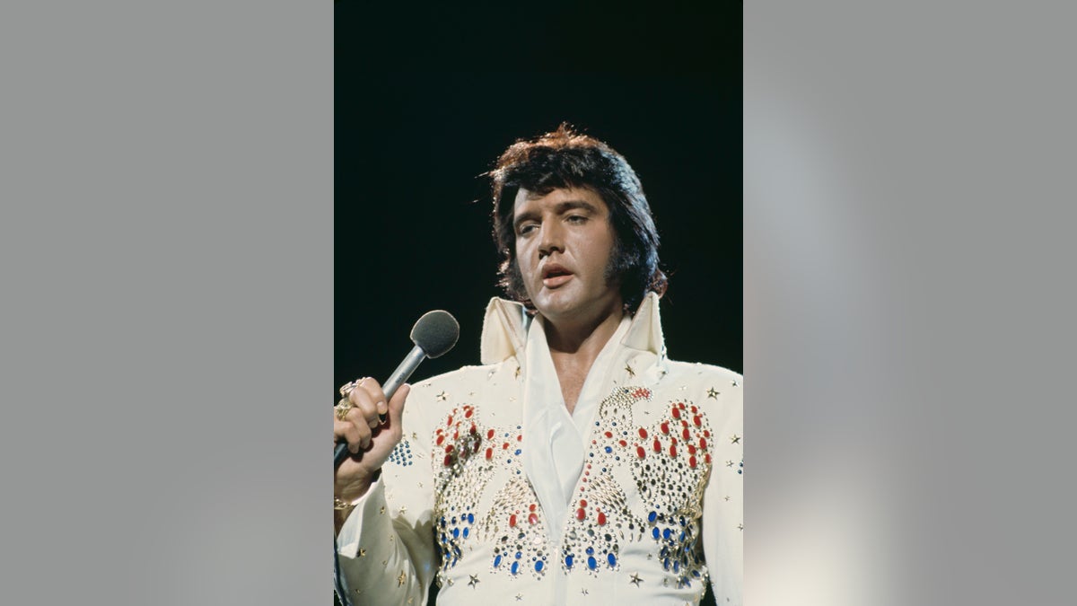 Um close de Elvis Presley vestindo um macacão branco enfeitado com joias no palco segurando um microfone