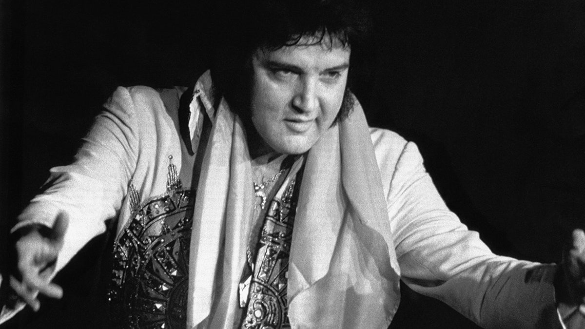 Um close de Elvis Presley no palco em um macacão branco com os braços da esposa abertos