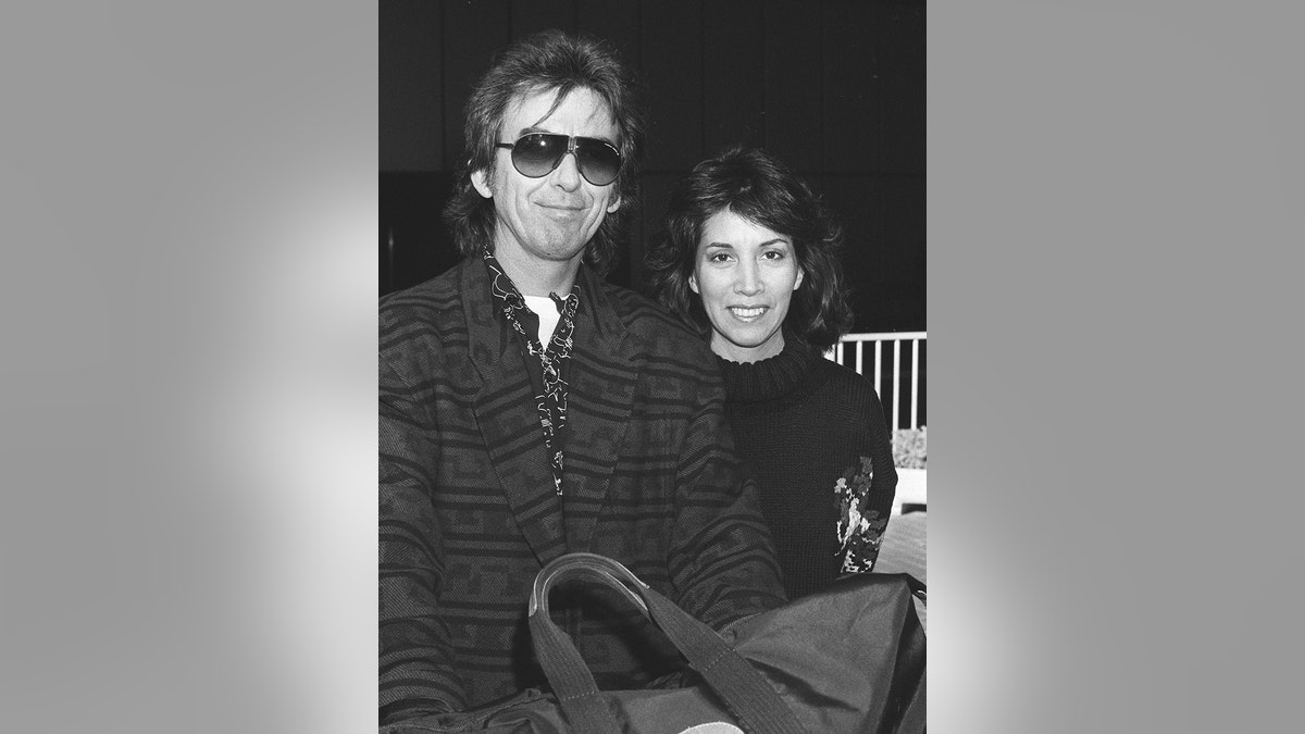 George Harrison smiling and wearing sunglasses and a dark coat next to his wife Olivia