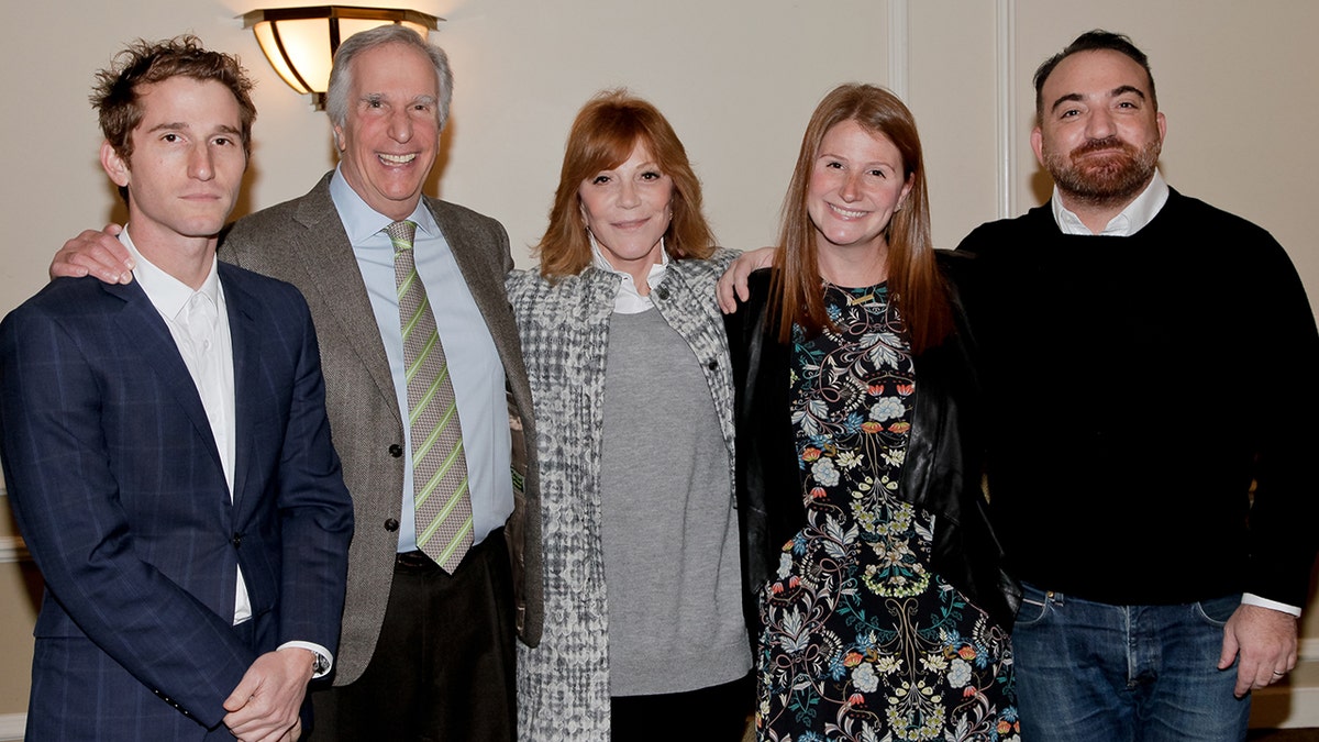 Henry Winkler in a grey suit, a blue shirt and a green striped tie surrounded by his children and their wife in outfits of similar colors