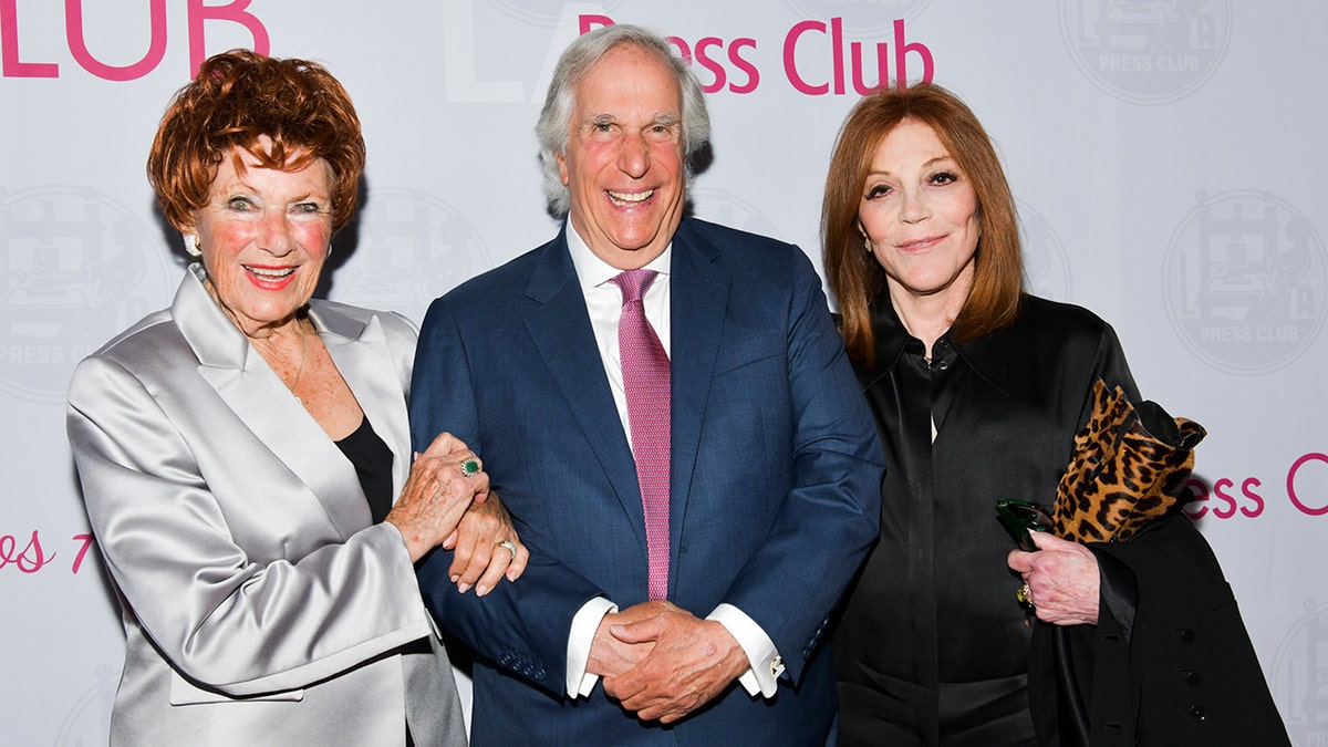 Henry Winkler smiling in a blue suit and pink tie next to Marion Ross in a silver suit and black shirt and his wife Stacey in a black dress