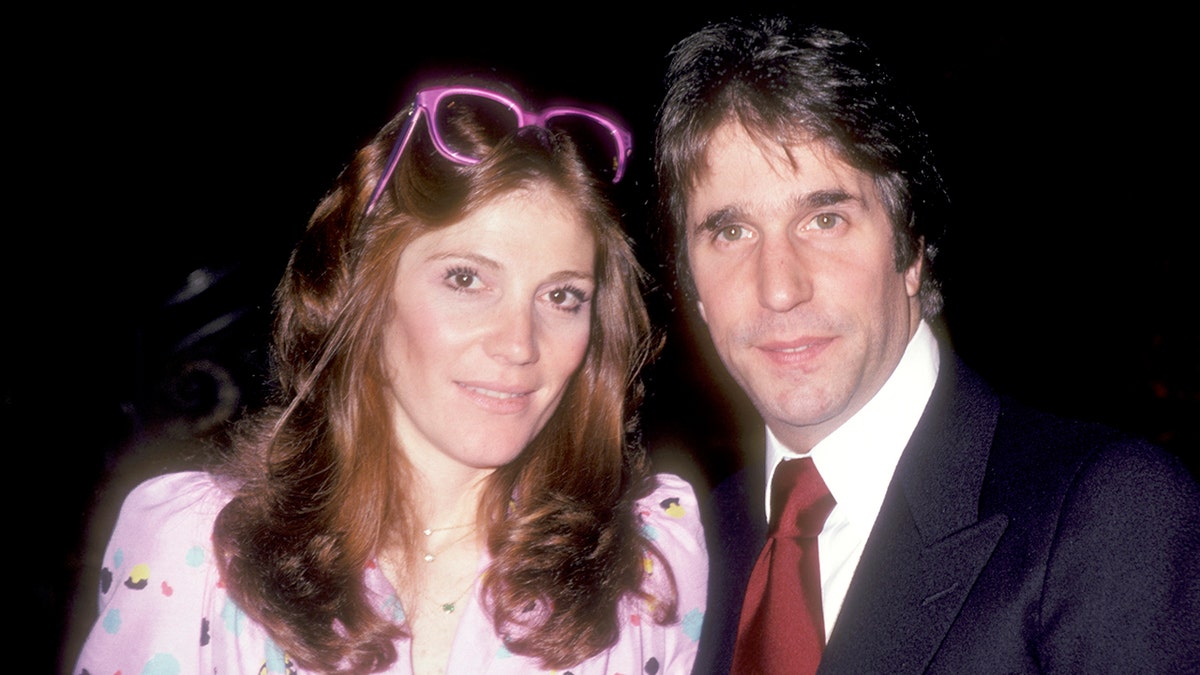 A close-up of Henry Winkler wearing a suit and a burgundy tie next to Stacey in a pink dress and purple sunglasses on her head