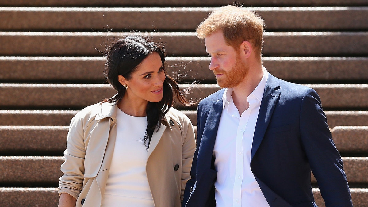 Meghan Markle wearing a white dress and a beige trench coat next to Prince Harry in a blue blazer and white shirt