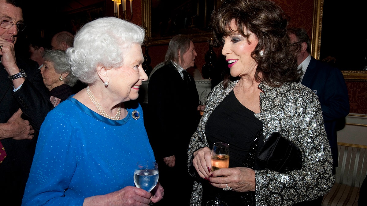 Queen Elizabeth wearing blue and holding a glass as she smiles at Joan Collins wearing a black dress, a silver blazer and holding a wine glass