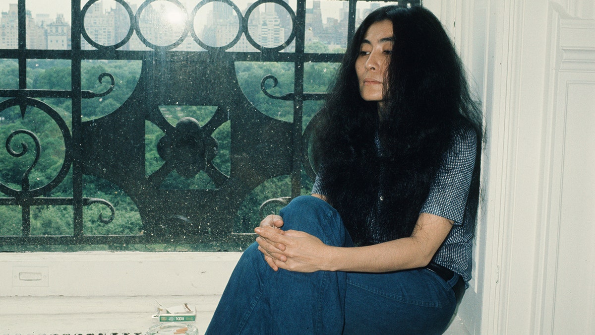 Yoko Ono looking somber sitting in front of a window at The Dakota
