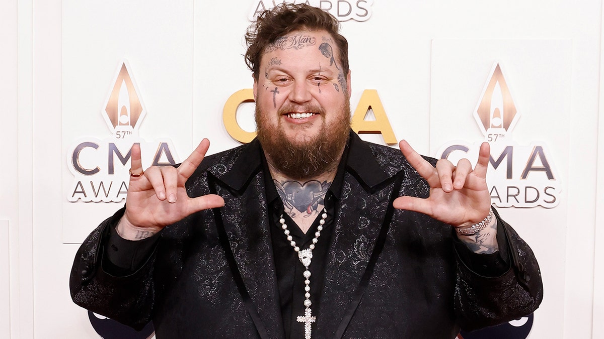 Jelly Roll in a black jacket at the CMAS puts up his two hands and does sign language for 'I love you'