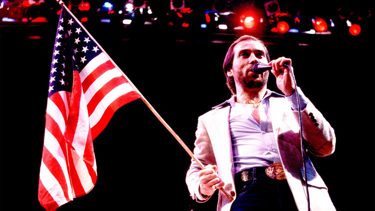 Lee Greenwood in 1984 holding US flag and microphone