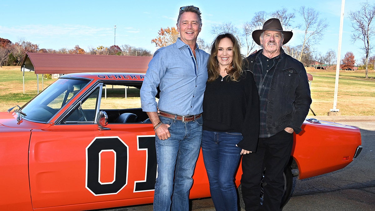 John Schneider, Catherine Bach and Tom Wopat pose close to each other in front of a red car outdoors