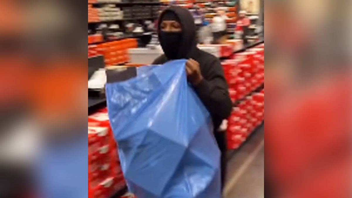 Person with mask over face, dressed in black, with blue trash bag stealing from Nike store