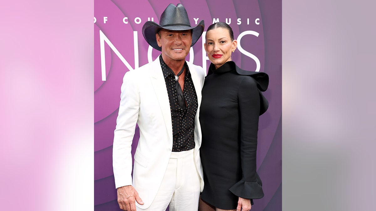 Tim McGraw in a white jacket and black shirt and black cowboy hat poses with Faith Hill in a black ruffle dress on the carpet